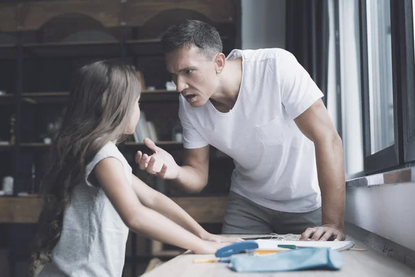El padre con una camiseta blanca le grita a su pequeña hija morena, que está junto a él cerca de la mesa de pino —  Fotos de Stock
