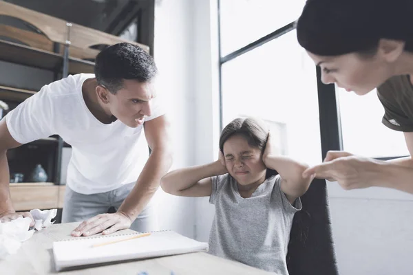 Um homem e uma mulher gritam furiosamente com uma menina de cabelos escuros que se senta em uma mesa perto da janela, com as mãos sobre as orelhas — Fotografia de Stock