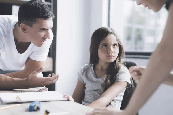 Un hombre y una mujer regañan a una niña que se sienta con un escritorio. La chica mira furiosamente a la mujer — Foto de Stock