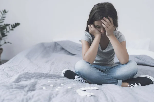 The girl sits on the bed with her head in her hands and looks desperately at the white pills that lie before her — Stock Photo, Image