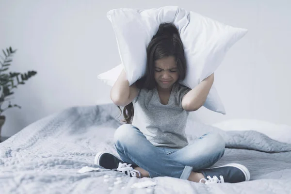 Het meisje zit met gekruiste benen op het bed, die betrekking hebben op haar hoofd met een kussen en fronsen, voor haar leugen tabletten — Stockfoto