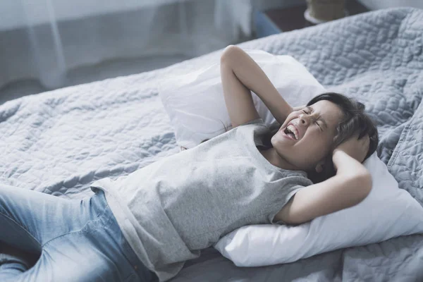 O fetiță țipă de durere din cap cu mâinile și se sprijină pe perna albă care se află pe pat. — Fotografie, imagine de stoc