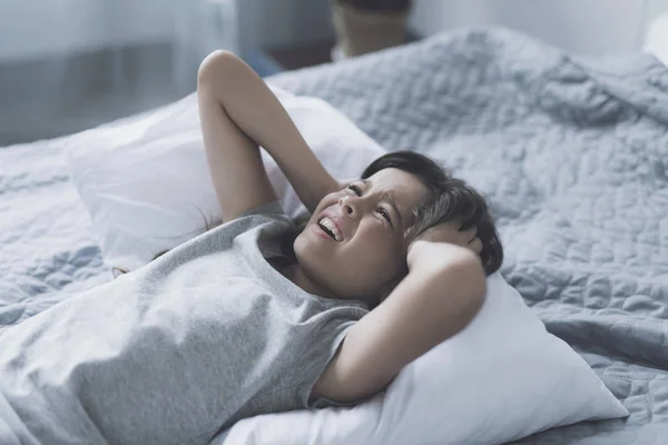 Una niña pequeña está acostada en una cama gris, apoyada en una almohada blanca y abrazando su cabeza en sus manos con dolor —  Fotos de Stock