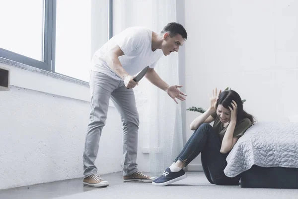 Un hombre con un cuchillo en las manos le grita a su esposa, que se sienta al lado del suelo con temor de llorar y cubrirse —  Fotos de Stock