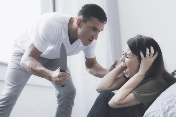 A woman screams with fear with her hands behind her head while she is threatened by an evil man with a knife — Stock Photo, Image