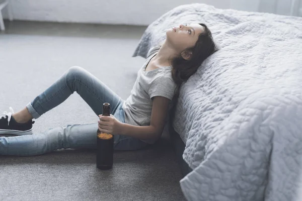 Chica sentada en el suelo apoyada en una cama gris y mirando al techo, sosteniendo en su mano una botella abierta de cerveza — Foto de Stock