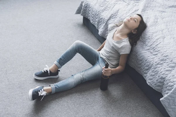 Темноволоса дівчина заснула, сидячи на підлозі, її голова на ліжку з пляшкою пива в руках — стокове фото
