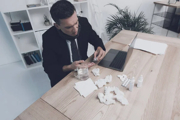 Хвора людина сидить на своєму робочому місці в офісі. У нього в руках пігулка і склянка води — стокове фото