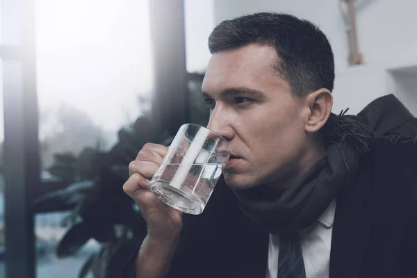 Nemocný muž sedí na jeho pracovišti v kanceláři. V rukou drží šálek vody — Stock fotografie