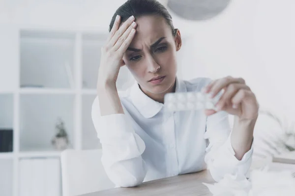 Kranke Frau sitzt an ihrem Arbeitsplatz im Büro. Sie hält ein Päckchen Tabletten in der Hand — Stockfoto