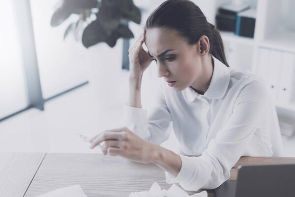 Больная женщина сидит на своем рабочем месте в офисе. У неё в руках пакет таблеток.
