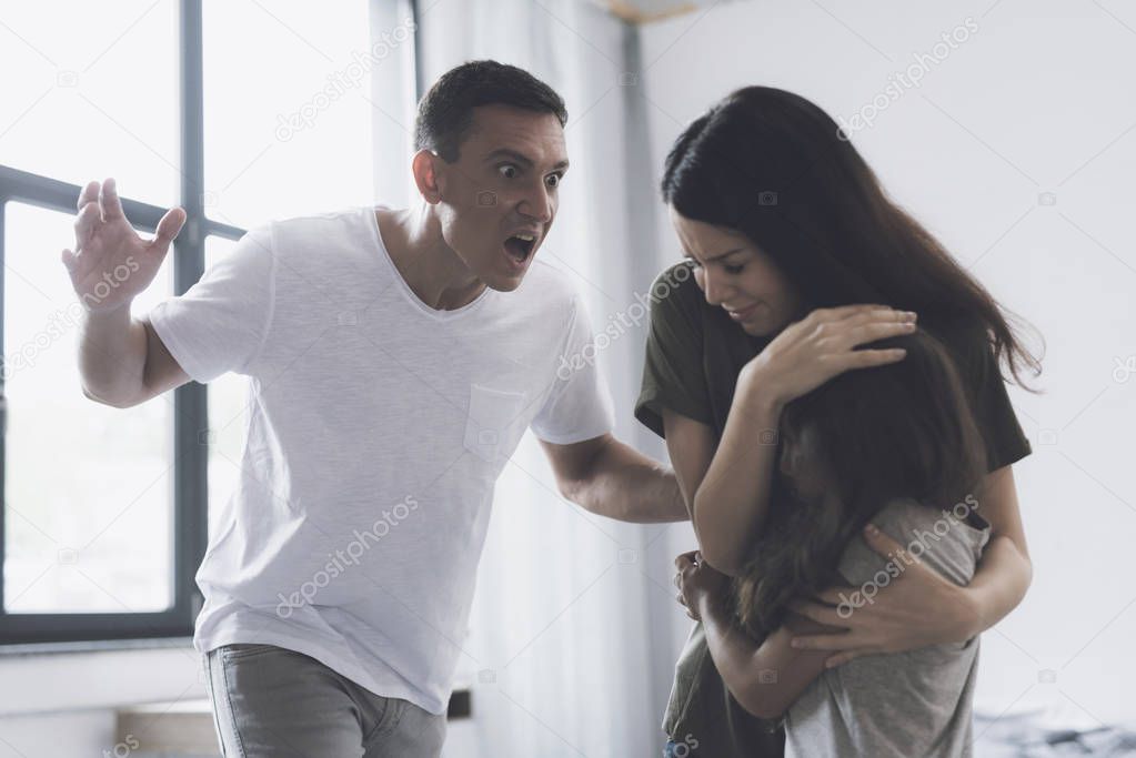 Wife and daughter are embracing, while her husband angrily screams at them and swings to strike