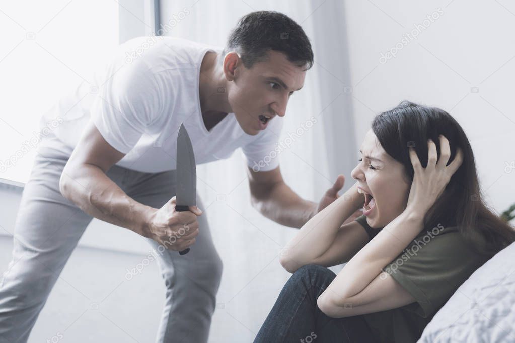 A woman screams with fear with her hands behind her head while she is threatened by an evil man with a knife