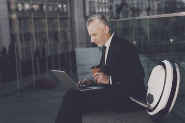 Bir adam bir parapetin üzerinde oturan bir dizüstü bilgisayar üzerinde çalışıyor. Onun mono-wheel