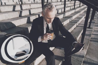 Sıkı iş takım elbiseli saygın bir yaşlı adam office merdivenlerinde oturur. O onun mono-tekerlek yanındaki bir tablet üzerinde çalışıyor