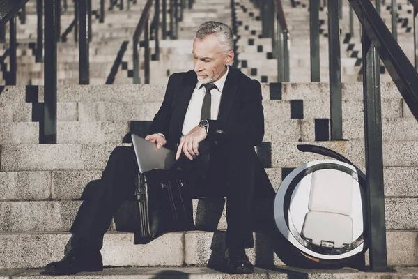 Ein respektabler alter Mann im strengen Businessanzug sitzt auf den Stufen des Büros. er arbeitet an einem Tablet, neben seinem Monorad — Stockfoto