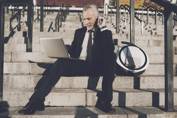 Un anciano respetable con un traje de negocios estricto se sienta en los escalones de la oficina. Trabaja en una tableta, junto a su monorueda. — Foto de Stock