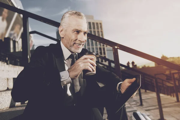 A respectable old man in a strict business suit sits on steps of office. He works on a tablet, next to his mono-wheel — Stock Photo, Image