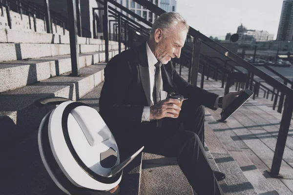 Un anciano respetable con un traje de negocios estricto se sienta en los escalones de la oficina. Trabaja en una tableta, junto a su monorueda. — Foto de Stock