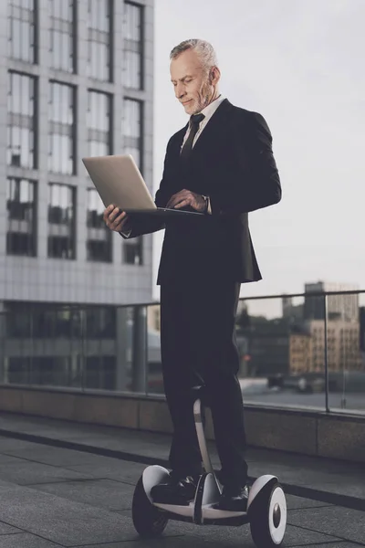 The old man is riding a gyroboard and is holding an open laptop in his hands. He has a strict business suit