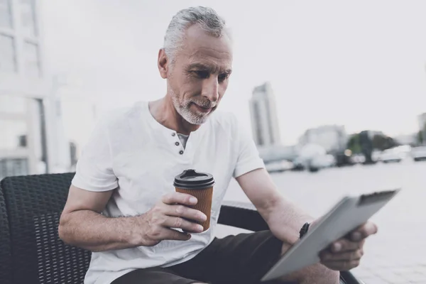 Un anciano con barba se sienta en un sofá en la calle. Él está sosteniendo café y una tableta — Foto de Stock