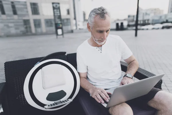 An old man with a beard sits on a sofa in the street. He is holding a laptop in his hands. Near his gyroboard