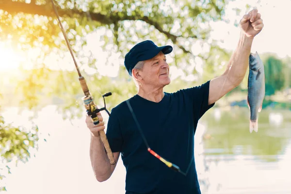 The old man in a black T-shirt is standing on the river bank, holding a pinning and looking at the fish he just caught — Stock Photo, Image