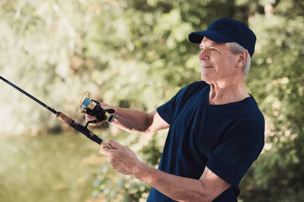 An elderly man stands on the river bank with a spinning in his hands, turns the fishing reel and stares at the water — Stock Photo, Image