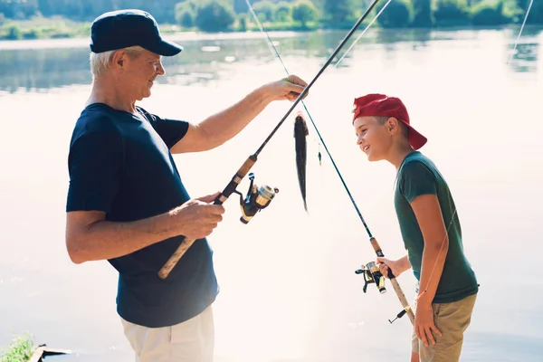 Stařec a chlapec stojí na břehu řeky s rotující zbraní. Starý muž ukazuje chlapce ryba, kterou chytil — Stock fotografie
