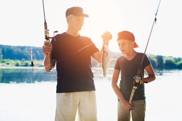 Avô e seu neto ficar na margem do rio com varas de fiação e olhar para o peixe que o velho pegou — Fotografia de Stock