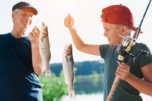 A boy with a spinning in his hands shows the old man what fish he caught. The old man keeps the same fish in his hands — Stock Photo, Image