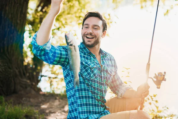 Ein Mann im blauen Hemd sitzt mit einer Spinnrute am Ufer des Flusses und freut sich über den Fisch, den er gerade gefangen hat — Stockfoto