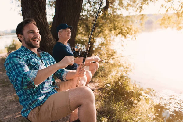 A man and an old man on the river bank. The man laughs and prepares to pull out the fish, the old man quietly fishes — Stock Photo, Image