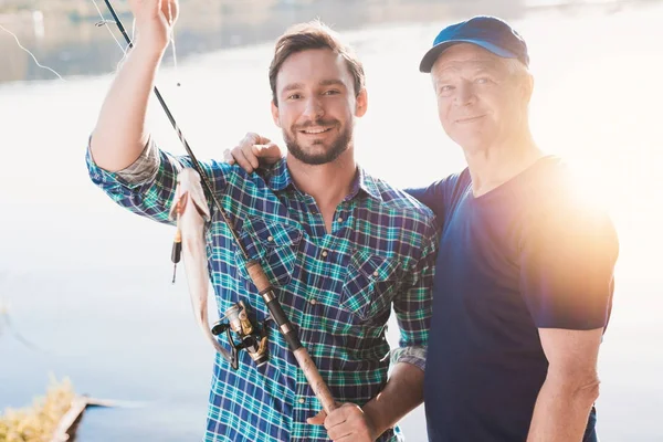 A man and an old man are posing with a fish that a man caught. Behind them, the sun is reflected in the river