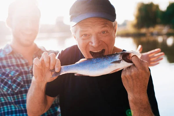 Um velho com uma t-shirt preta a posar enquanto segura um peixe. Ele finge que quer comê-la — Fotografia de Stock