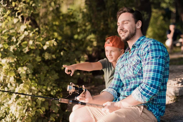 A man with his son sits on the river bank and catches fish. The boy shows him something on the river — Stock Photo, Image