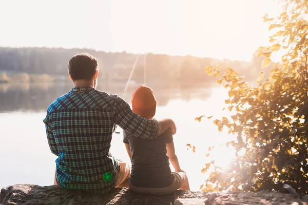 Padre e hijo están pescando, sentados en la orilla del río contra el telón de fondo del amanecer — Foto de Stock