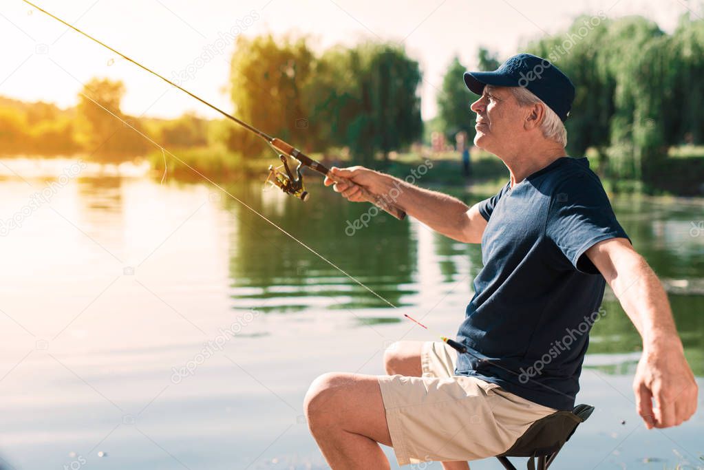 The old man is sitting on the river bank with a spinning in his hands and preparing to throw in an ode fish hook