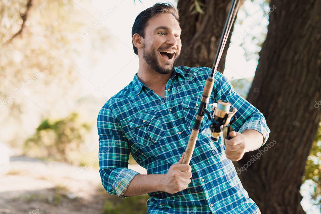 A happy man in a blue shirt stands on the river bank and holds a spinning in his right hand, twisting reel by left