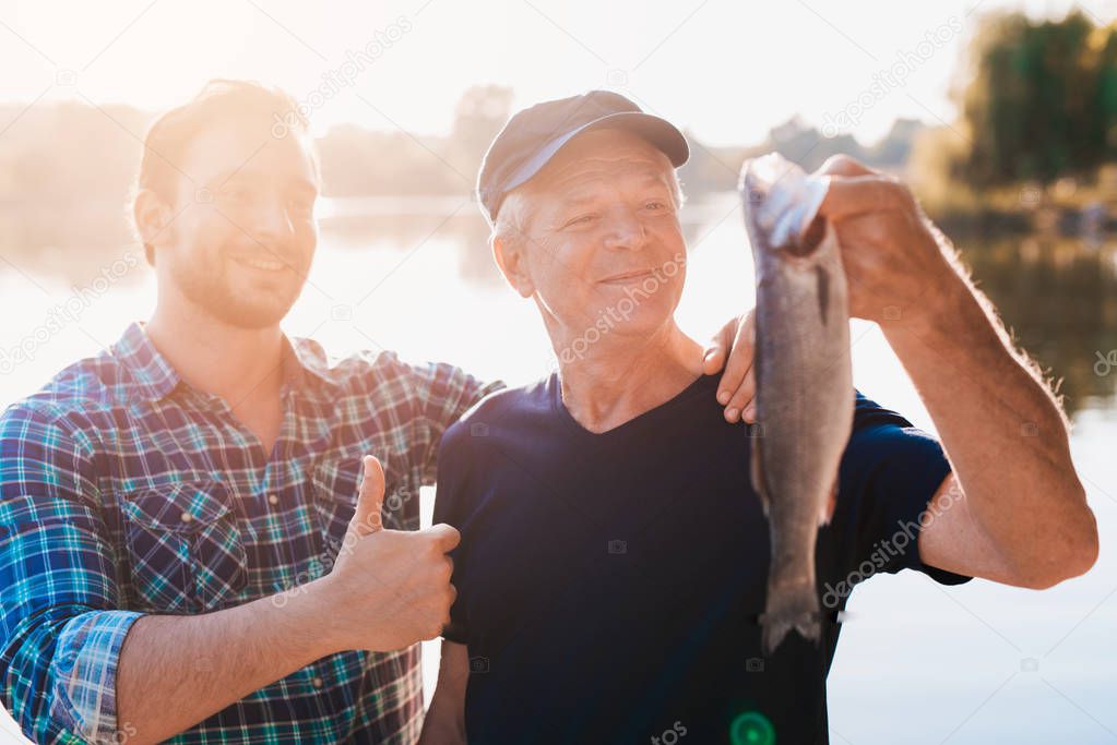Thumbs up. The old man is holding a fish. A man stands behind him with his thumb up