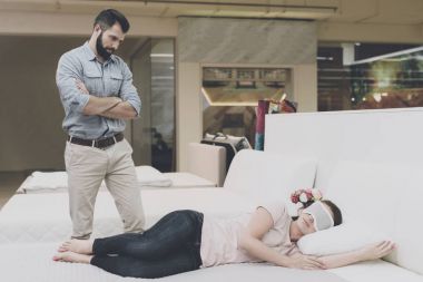 The woman put on a blindfold on her eyes and fell asleep on the bed in the store. A man looks at her with displeasure clipart