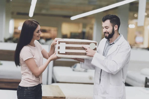 Consultor-ortopedista ayuda a la chica a elegir un colchón en la tienda. Él le ofrece un colchón y muestra su muestra — Foto de Stock