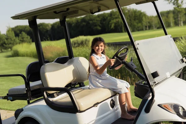 En liten flicka sitter barfota i en vit golfbil och hängande bakom ratten — Stockfoto