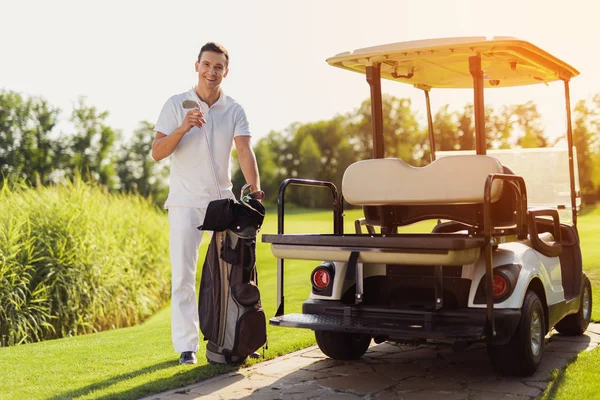 Un hombre sonriente con un traje blanco sostiene un palo de golf junto a un carrito de golf —  Fotos de Stock