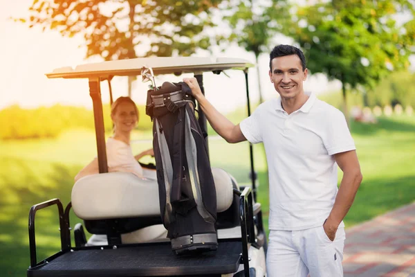 Una mujer se sienta detrás del volante de un carrito de golf, un hombre está en primer plano y sostiene una bolsa con palos de golf —  Fotos de Stock