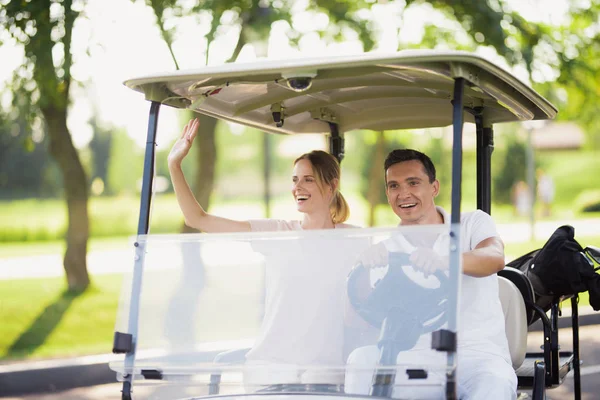 Ett par sitter i en vit golfbil. En man kör, en kvinna sitter och välkomnar någon — Stockfoto
