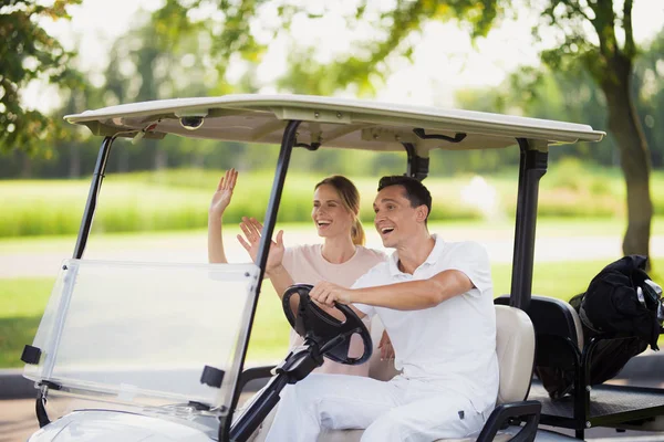 Un hombre y una mujer están sentados en un carro de golf blanco y saludando a alguien delante de ellos —  Fotos de Stock