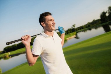 Beyaz elbiseli bir adam bir golf sahasında ayakta ve gülümseyerek, bir golf kulübü arkasında holding