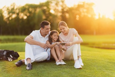 The family rests after playing golf and look at the smartphone screen together. They are happy and smiling