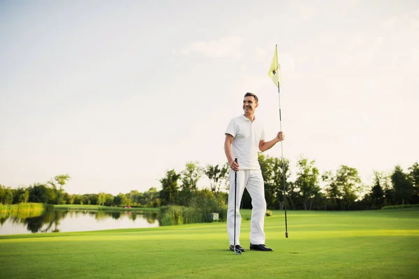 A man stands on a golf course, holds a golf club and flags for the hole
